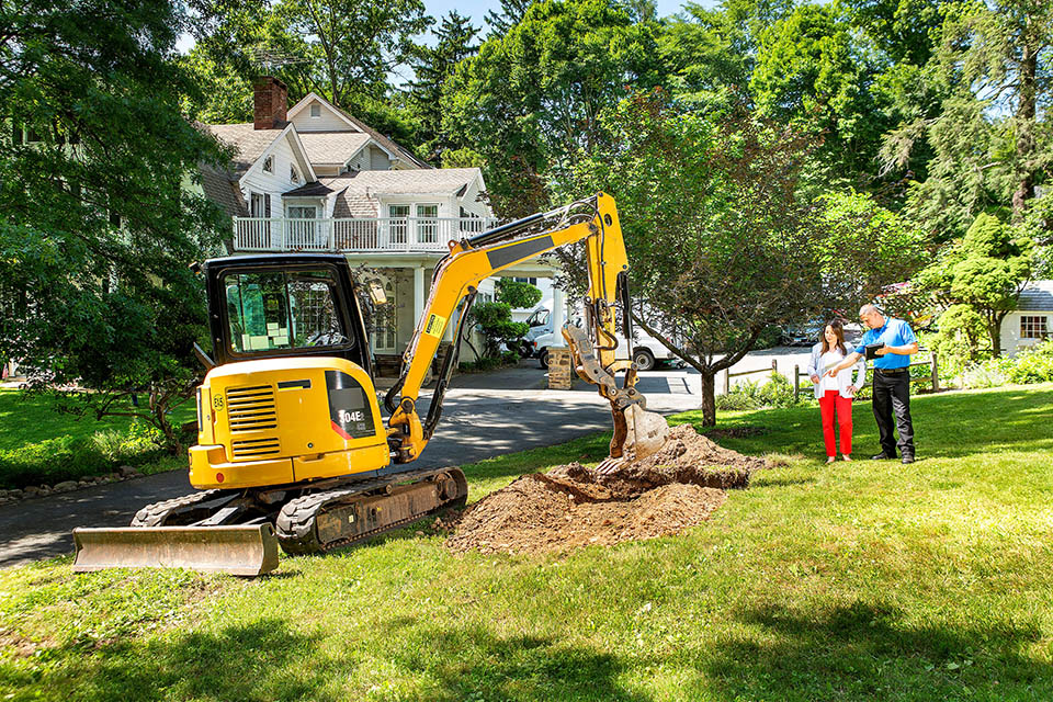 HVAC technician and homeowner standing next to bulldozer in front yard as geothermal installation is being performed
